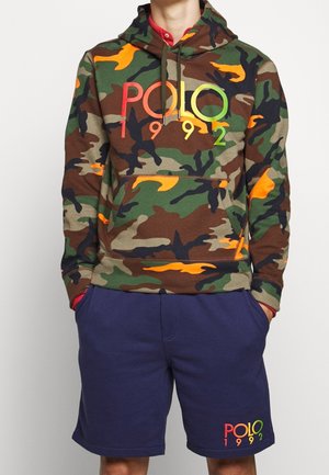 Mężczyzna ubrany w zieloną, brązową i pomarańczową kamuflażową bluzę z kapturem z napisem "POLO 1992", w połączeniu z granatowymi szortami również oznaczonymi "POLO 1992".