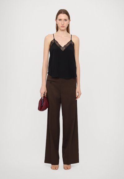 Zwarte cami met kantdetails, gecombineerd met brede bruine broeken. Model houdt een bordeauxrood tasje vast, staande voor een lichtgrijze achtergrond.