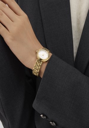 Kuldne kell, millel on ümmargune korpus, valge sihverplaat ja teemantilaadsed tunni näitajad, millel on tekstuuriga kuldne metallist randmeosa. Kandmisel halli ülikonna varrukas.