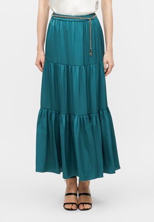 Femme portant une jupe maxi échelonnée couleur sarcelle avec une ceinture chaîne dorée, associée à des sandales noires à bout ouvert et un haut blanc sans manches.