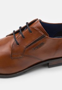 Zapato de cuero marrón con superficie lisa, cordones negros, perfil delgado y un pequeño logotipo de la marca en el lateral. Un diseño discreto.