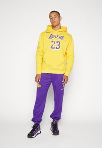 Moletom amarelo com "Lakers 23" em roxo, combinado com calças de moletom roxas com o logótipo dos Lakers. Ténis pretos com detalhes em roxo.
