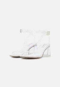 Sandales à talons hauts blanches avec des brides transparentes et des talons carrés transparents. Avec une bride de cheville à boucle et un coussin de pied lisse.