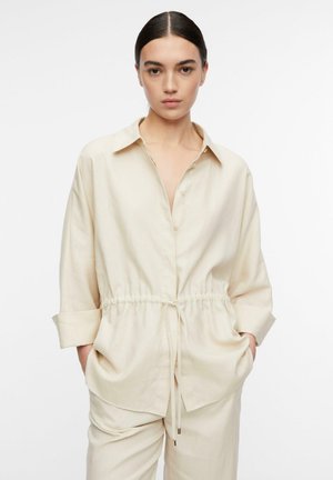 Frau mit zurückgebundenen dunklen Haaren, trägt ein lockeres beiges Hemd mit Knöpfen und Kordelzug in der Taille und passende beigen Hosen, Hände in den Taschen.
