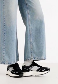 Zapatillas en blanco y negro con parte superior texturizada, diseño de cordones y una suela blanca gruesa. Combinadas con unos vaqueros de tiro alto y pierna ancha en azul claro.