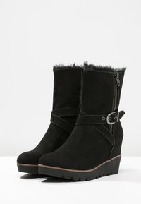 Bottines noires à talon compensé avec fermeture éclair latérale, bride à boucle, doublure en fausse fourrure et semelle rainurée, présentées sur fond blanc.