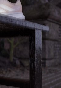 Weißer Sneaker, der auf die Kante eines nassen, dunklen Holztisches tritt, mit einer unscharfen Steinmauer und einem Außenhintergrund.