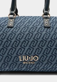 Denim-Handtasche mit einem sich wiederholenden geometrischen Muster in Dunkelblau, schwarzen Lederelementen, goldfarbenen Beschlägen und einem auffälligen Logo.