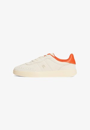 Zapatilla beige con superficie lisa, acento en el talón naranja, punta redondeada y suela de goma plana. Presenta una parte delantera con cordones y perforaciones sutiles.