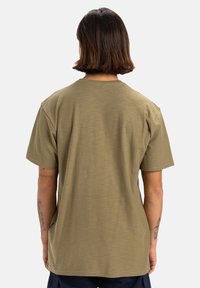 Camiseta de manga corta verde oliva con cuello redondo, corte relajado y tejido texturizado. Etiqueta visible en la costura lateral izquierda.