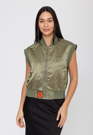 Femme aux longs cheveux bruns portant une veste olive verte sans manches à fermeture éclair et une jupe noire, debout devant un fond gris clair.