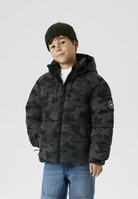 Veste doudoune noire camouflage avec capuche, fermeture éclair et poches latérales. Bonnet vert à côtes. Jean bleu à coupe relax. 
