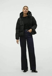 Kvinde står iført en sort hættejakke, sort top, mørke brede bukser og sorte hæle støvler mod en hvid baggrund.