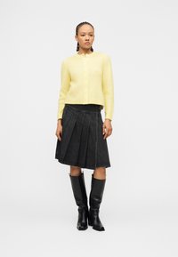 Γυναίκα με πλεκτά μαλλιά που φοράει κίτρινη ζακέτα με κουμπιά, μαύρη πλισέ φούστα και μαύρες δερμάτινες μπότες έως το γόνατο, στέκεται μπροστά σε ένα λευκό φόντο.