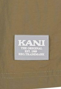Tissu vert olive avec une étiquette rectangulaire grise comportant un texte blanc : "KANI THE ORIGINAL EST. 1989 REG. TRADEMARK," cousu.