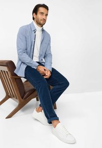 Lichtblauw overhemd met knopen boven een witte coltrui, gecombineerd met marineblauwe corduroypantalons en witte sneakers, zittend op een houten stoel.