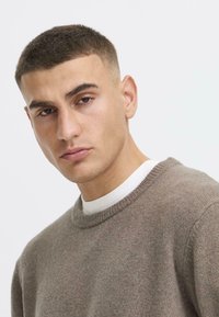 Brown knitted sweater with a ribbed neckline, worn over a white shirt. The material appears soft and textured, with a casual fit.