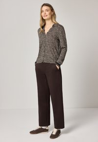 Femme debout les mains dans les poches, portant une chemise à manches longues imprimée léopard marron, un pantalon noir et des baskets marron à lacets blancs.