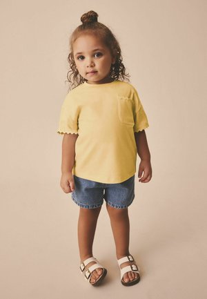 Väikelaps lokkis juustega, kollase särgi, teksapükste ja valgete sandaalidega seisab ühevärvilise beeži tausta ees.