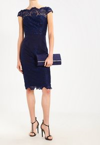 Tummansininen pitsimekko, jossa on hihat, kapea vartaloosa ja aaltoileva helma. Yhdistettynä sopivaan laukkuun ja mustiin sandaaleihin.