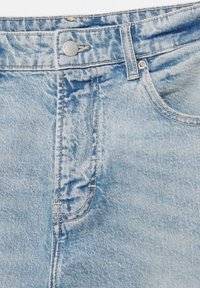 Detailný záber na svetlomodré džínsy ukazujúci predné vrecko, očko na opasok, kovové gombíky a švy na zapínaní.