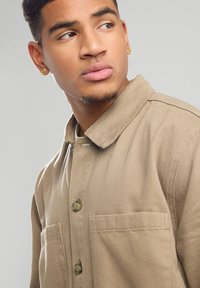 Camicia button-up color beige realizzata in tessuto testurizzato, con colletto e due tasche frontali. Il modello ha i capelli corti e indossa piccoli orecchini d'oro.