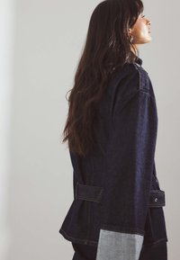 Woman with long dark hair wearing a dark denim jacket with wide cuffs, standing in profile against a plain light wall.