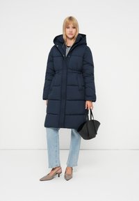 Palton puffer bleu marin cu glugă, talie strânsă, buzunare mari și nasturi în față, asortat cu blugi bleu deschis și o geantă neagră.