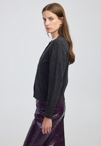 Vrouw met lang bruin haar, goudkleurige oorbellen, een donkergrijze cardigan en een paarse leren rok, staande in profiel tegen een effen achtergrond.