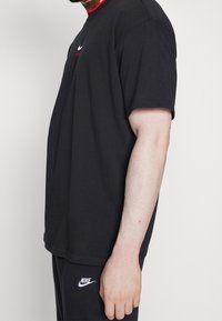 Man wearing black Nike t-shirt and pants, showing side profile of upper body and arm against plain background.