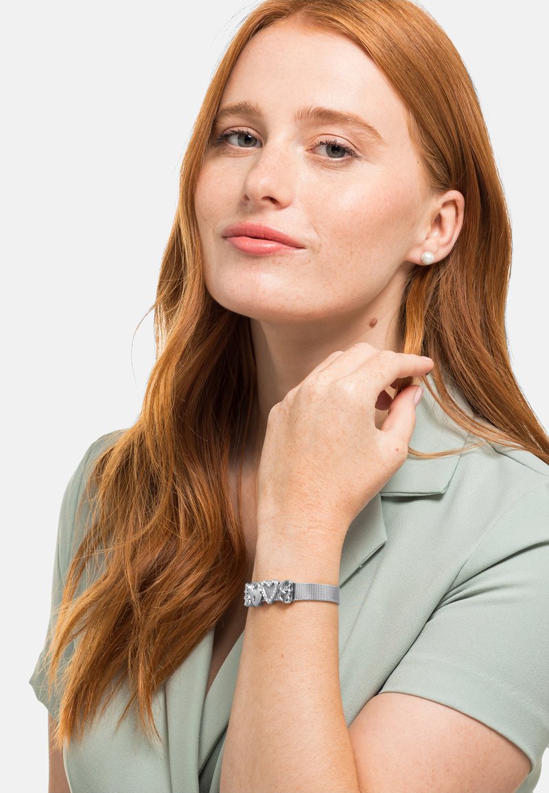 Bracciale in rete argentata con accenti metallici decorati, indossato su un polso nudo. La mano tocca delicatamente il collo, sullo sfondo chiaro.