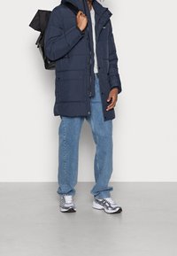 Granatowa kurtka puchowa z kapturem, zamkiem błyskawicznym i przednimi kieszeniami, w połączeniu z jasnoniebieskimi dżinsami i srebrnymi butami do biegania. Plecak na ramieniu.