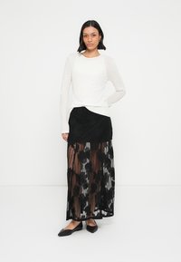 Longue jupe en dentelle noire avec motif floral, superposée à un jupon noir uni. Haut blanc à manches longues, coupe ajustée. Ballerines noires.