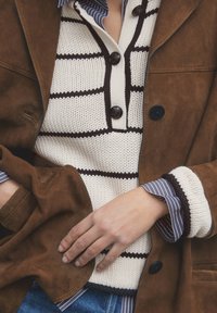 Veste en daim marron superposée sur un pull crème avec des rayures marron et une patte de boutonnage. Les poignets d'une chemise à rayures bleues et blanches sont visibles.