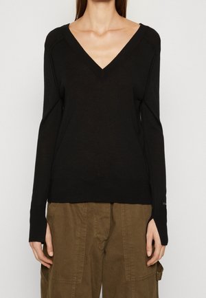 Femme portant un pull noir à manches longues col en V et un pantalon marron ample, debout les bras détendus le long du corps sur un fond uni.