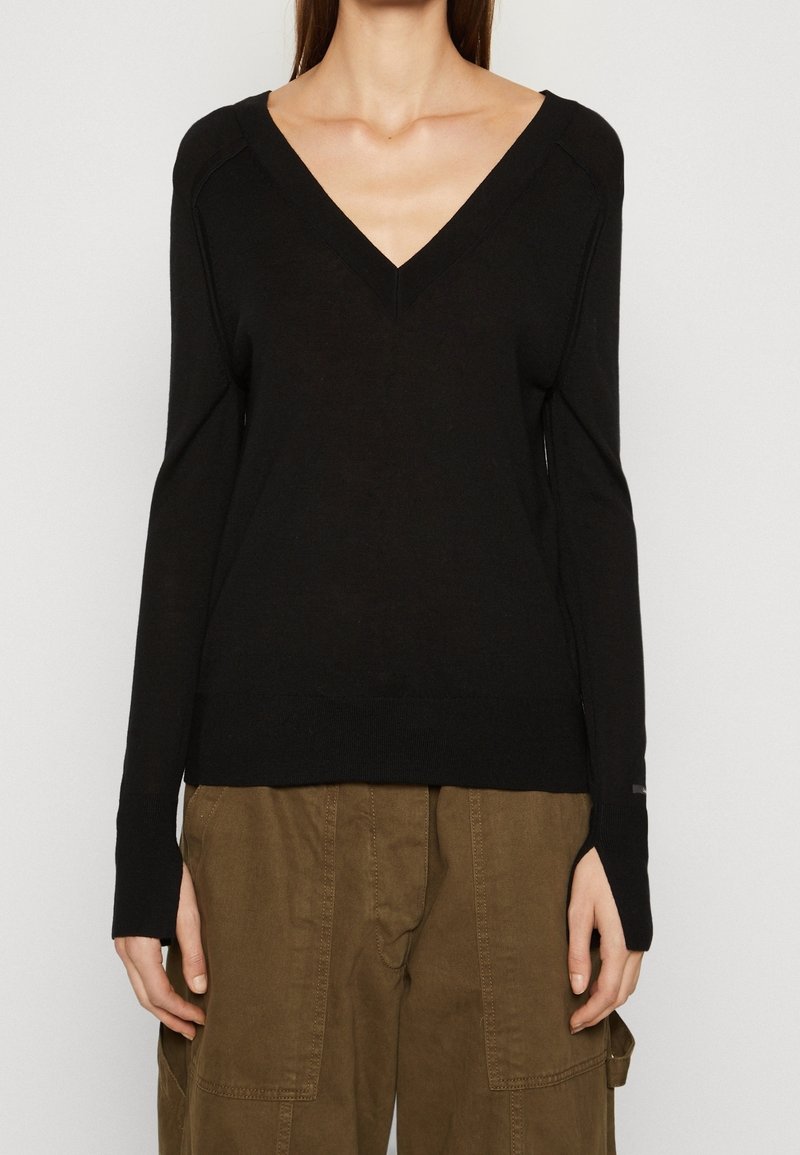 Femme portant un pull noir à manches longues col en V et un pantalon marron ample, debout les bras détendus le long du corps sur un fond uni.