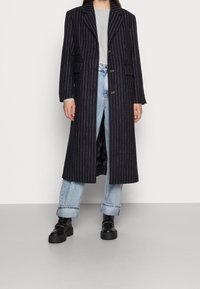 Manteau long et ajusté en marine à fines rayures blanches. Il présente un col cranté, deux boutons et des poches avant. Porté avec un jean bleu clair et des bottes.