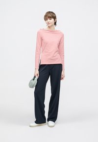 Czerwono-biała bluzka w paski z długim rękawem i dekoltem w łódkę, zestawiona z ciemnoniebieskimi szerokimi spodniami oraz białymi tenisówkami. Trzyma zieloną torbę.