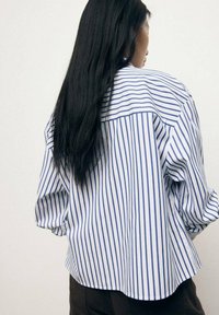 Blue and white striped oversized shirt with a loose fit, featuring a curved hem and rolled sleeves, made from lightweight fabric.