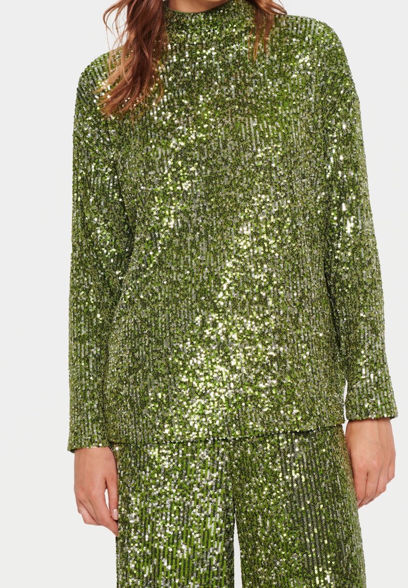 Donna che indossa un top a maniche lunghe e pantaloni abbinati verdi con paillettes e motivi verticali scintillanti su uno sfondo semplice.