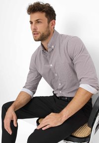 Camicia a quadri rossi e bianchi con colletto abbottonato; maniche arrotolate con polsini bianchi. Seduto su una sedia, indossa pantaloni neri.