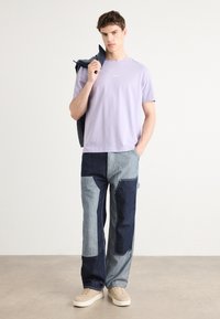 Hellblaues Baumwoll-T-Shirt mit einem Logo, kombiniert mit zweifarbigen Patchwork-Jeans in Dunkel- und Hellblau, ergänzt durch beigen Sneakers.