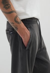 Graue Jogginghosen mit einem elastischen Bund und Kordelzug, seitlichen Taschen und einem strukturierten Stoff. Enthält ein kleines Markenschild an der linken Tasche.