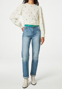 Maglione bianco con ricami floreali verdi, abbinato a jeans a gamba dritta azzurri e una cintura turchese. Stivali alla caviglia color crema.