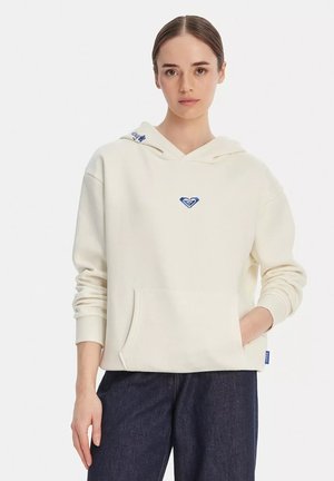 Donna che indossa una felpa bianco sporco con un piccolo logo a forma di cuore blu al centro e jeans blu scuro, in piedi con una mano in tasca.