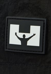 Schwarzer Textilhintergrund mit einem rechteckigen schwarz-weißen Patch. Der Patch zeigt die Silhouette einer Person mit erhobenen Armen.