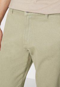 Pantaloni di cotone verde chiaro con una finitura testurizzata, dotati di tasche frontali e chiusura con bottone in vita. Design casual, vestibilità sartoriale.