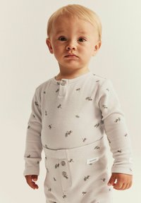 Un tout-petit aux cheveux clairs portant un pyjama blanc à manches longues en texture gaufrée avec de petites impressions de cerises, se tenant contre un fond clair uni.