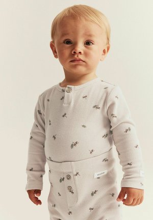Un tout-petit aux cheveux clairs portant un pyjama blanc à manches longues en texture gaufrée avec de petites impressions de cerises, se tenant contre un fond clair uni.