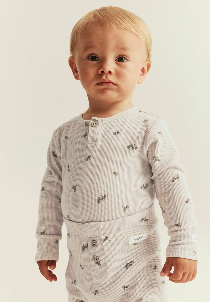 Un tout-petit aux cheveux clairs portant un pyjama blanc à manches longues en texture gaufrée avec de petites impressions de cerises, se tenant contre un fond clair uni.